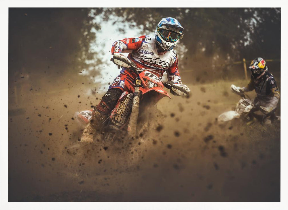 An action shot of a motocross rider in red, white, and blue gear, speeding through a dusty turn on a dirt track, kicking up a large cloud of mud and debris. Another racer is visible in the background. The image is presented within a silver frame. Print