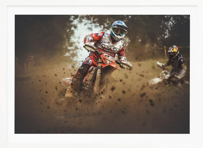An action shot of a motocross rider in red, white, and blue gear, speeding through a dusty turn on a dirt track, kicking up a large cloud of mud and debris. Another racer is visible in the background. The image is presented within a silver frame. Print