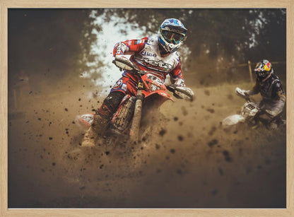 An action shot of a motocross rider in red, white, and blue gear, speeding through a dusty turn on a dirt track, kicking up a large cloud of mud and debris. Another racer is visible in the background. The image is presented within a silver frame. Print