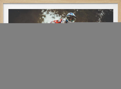 An action shot of a motocross rider in red, white, and blue gear, speeding through a dusty turn on a dirt track, kicking up a large cloud of mud and debris. Another racer is visible in the background. The image is presented within a silver frame. Print