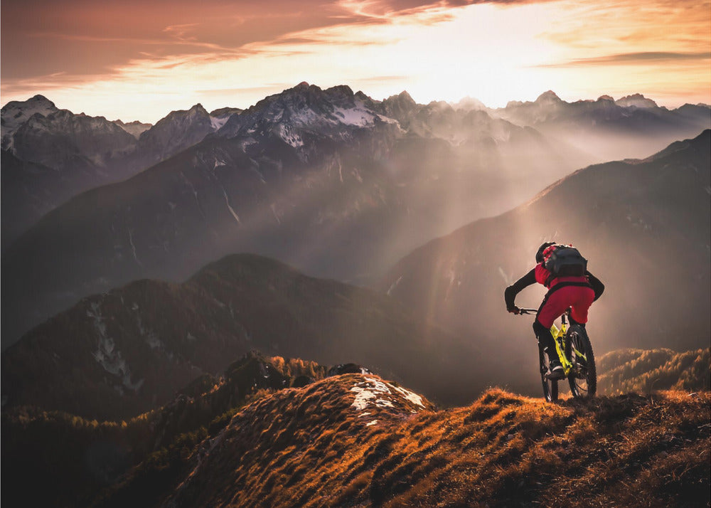 A mountain biker in red and black gear pauses on a grassy, sunlit ridgeline, looking out at a vast mountain range with snow-capped peaks as the sun sets behind them, casting dramatic rays of light through the valleys. Poster