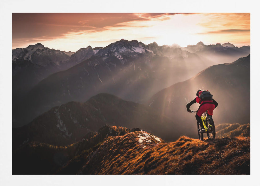 A mountain biker in red and black gear pauses on a grassy, sunlit ridgeline, looking out at a vast mountain range with snow-capped peaks as the sun sets behind them, casting dramatic rays of light through the valleys. Poster