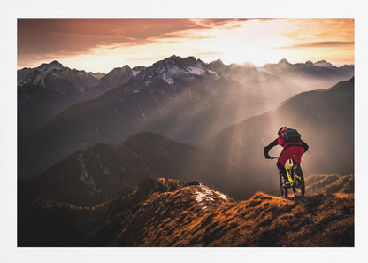 A mountain biker in red and black gear pauses on a grassy, sunlit ridgeline, looking out at a vast mountain range with snow-capped peaks as the sun sets behind them, casting dramatic rays of light through the valleys. Poster