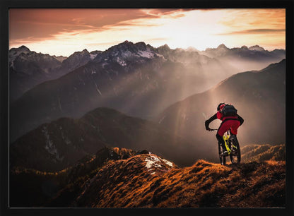 A mountain biker in red and black gear pauses on a grassy, sunlit ridgeline, looking out at a vast mountain range with snow-capped peaks as the sun sets behind them, casting dramatic rays of light through the valleys. Poster