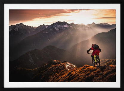 A mountain biker in red and black gear pauses on a grassy, sunlit ridgeline, looking out at a vast mountain range with snow-capped peaks as the sun sets behind them, casting dramatic rays of light through the valleys. Poster