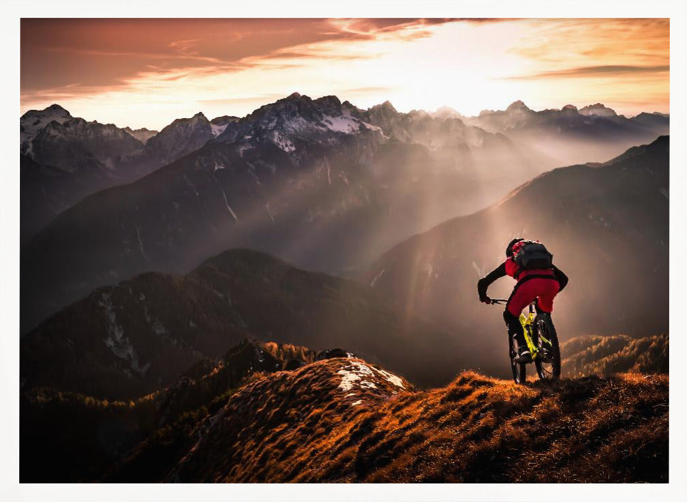 A mountain biker in red and black gear pauses on a grassy, sunlit ridgeline, looking out at a vast mountain range with snow-capped peaks as the sun sets behind them, casting dramatic rays of light through the valleys. Poster