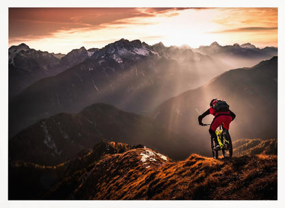 A mountain biker in red and black gear pauses on a grassy, sunlit ridgeline, looking out at a vast mountain range with snow-capped peaks as the sun sets behind them, casting dramatic rays of light through the valleys. Poster