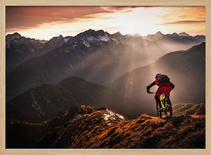 A mountain biker in red and black gear pauses on a grassy, sunlit ridgeline, looking out at a vast mountain range with snow-capped peaks as the sun sets behind them, casting dramatic rays of light through the valleys. Poster