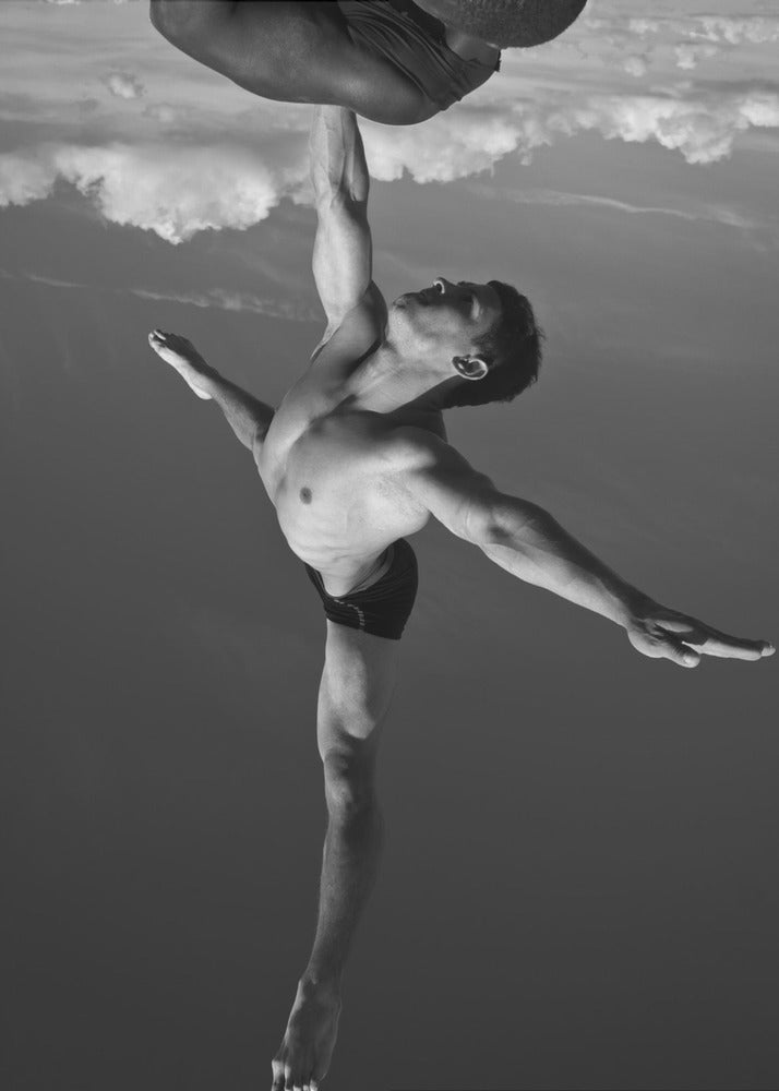 A surreal black and white photograph of two male acrobats. One man, in a graceful dance-like pose balancing on one leg, holds another man aloft with a single outstretched arm. The man being held is upside down against a backdrop of clouds, creating a gravity-defying and artistic illusion. Print