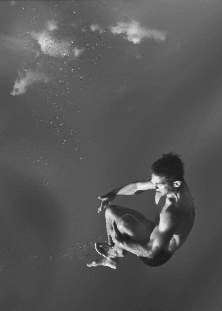 A dramatic black and white photograph of a male diver suspended mid-air in a tuck position. The background is a dark, atmospheric gray, with what appears to be clouds and bubbles, giving the impression of being underwater or in a vast, empty space. The lighting highlights the athlete's muscular physique. Artwork