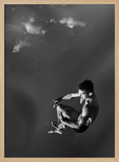 A dramatic black and white photograph of a male diver suspended mid-air in a tuck position. The background is a dark, atmospheric gray, with what appears to be clouds and bubbles, giving the impression of being underwater or in a vast, empty space. The lighting highlights the athlete's muscular physique. Artwork
