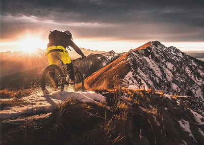 A mountain biker seen from behind rides along a narrow, snowy trail on a mountain ridge during a dramatic sunset, with the sun flaring brightly and a vast, snow-capped mountain range in the background. Decor