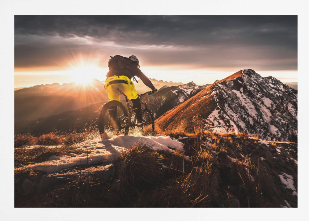 A mountain biker seen from behind rides along a narrow, snowy trail on a mountain ridge during a dramatic sunset, with the sun flaring brightly and a vast, snow-capped mountain range in the background. Decor