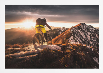 A mountain biker seen from behind rides along a narrow, snowy trail on a mountain ridge during a dramatic sunset, with the sun flaring brightly and a vast, snow-capped mountain range in the background. Decor