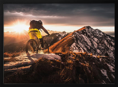 A mountain biker seen from behind rides along a narrow, snowy trail on a mountain ridge during a dramatic sunset, with the sun flaring brightly and a vast, snow-capped mountain range in the background. Decor
