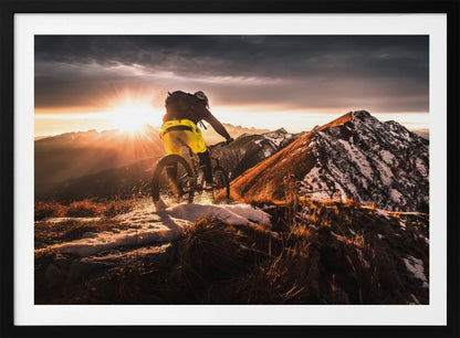 A mountain biker seen from behind rides along a narrow, snowy trail on a mountain ridge during a dramatic sunset, with the sun flaring brightly and a vast, snow-capped mountain range in the background. Decor
