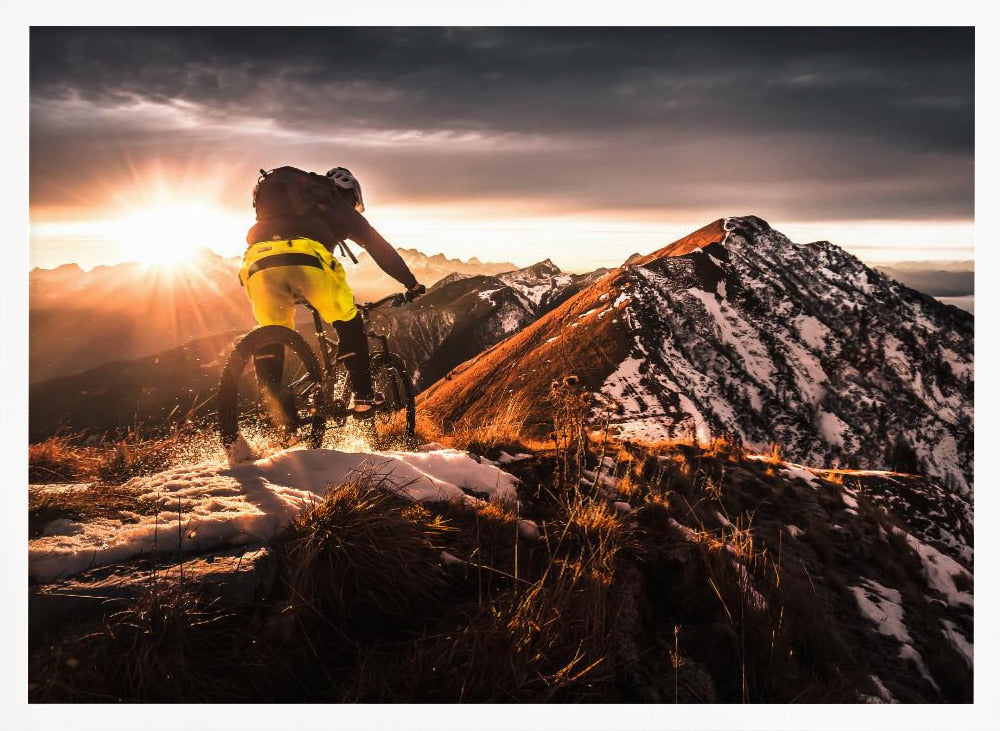A mountain biker seen from behind rides along a narrow, snowy trail on a mountain ridge during a dramatic sunset, with the sun flaring brightly and a vast, snow-capped mountain range in the background. Decor