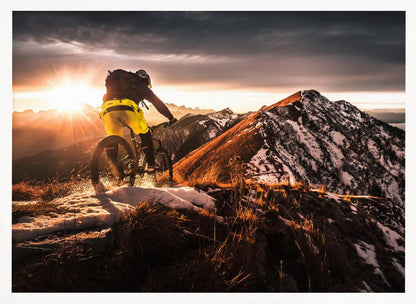 A mountain biker seen from behind rides along a narrow, snowy trail on a mountain ridge during a dramatic sunset, with the sun flaring brightly and a vast, snow-capped mountain range in the background. Decor