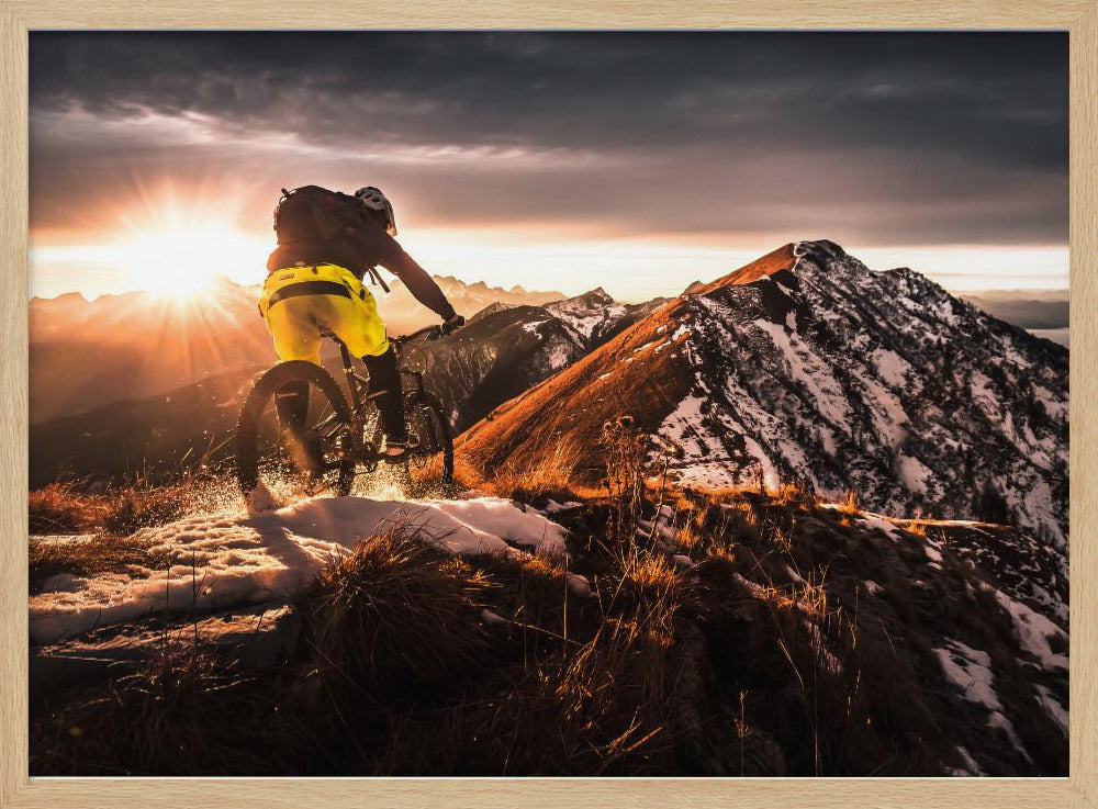 A mountain biker seen from behind rides along a narrow, snowy trail on a mountain ridge during a dramatic sunset, with the sun flaring brightly and a vast, snow-capped mountain range in the background. Decor