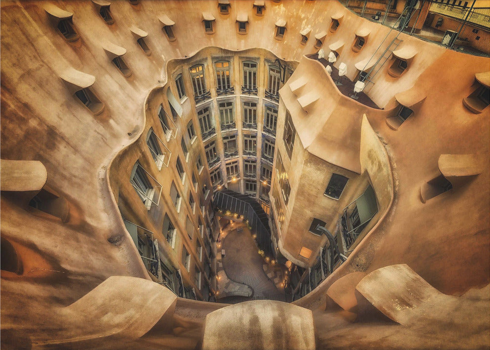 A high-angle, downward view of the inner courtyard of an architecturally unique building, likely Casa Milà by Gaudí. The warm, sandy-colored walls curve and undulate organically, surrounding the multi-story structure with its many windows. The courtyard below is lit, casting a warm glow. Decor
