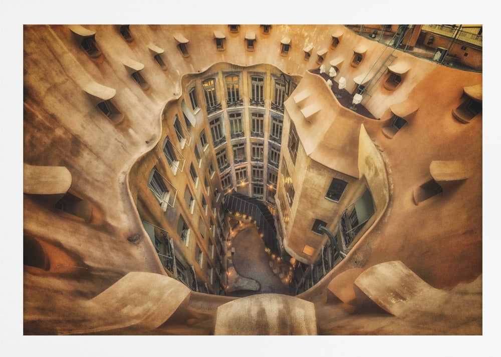 A high-angle, downward view of the inner courtyard of an architecturally unique building, likely Casa Milà by Gaudí. The warm, sandy-colored walls curve and undulate organically, surrounding the multi-story structure with its many windows. The courtyard below is lit, casting a warm glow. Decor