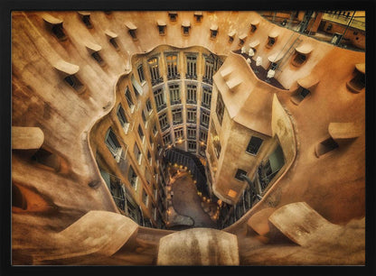 A high-angle, downward view of the inner courtyard of an architecturally unique building, likely Casa Milà by Gaudí. The warm, sandy-colored walls curve and undulate organically, surrounding the multi-story structure with its many windows. The courtyard below is lit, casting a warm glow. Decor