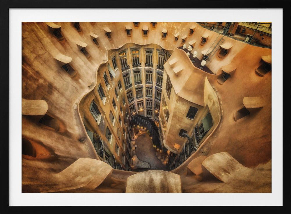 A high-angle, downward view of the inner courtyard of an architecturally unique building, likely Casa Milà by Gaudí. The warm, sandy-colored walls curve and undulate organically, surrounding the multi-story structure with its many windows. The courtyard below is lit, casting a warm glow. Decor