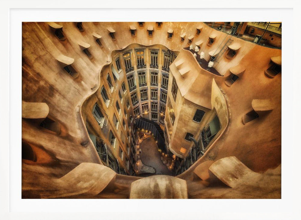 A high-angle, downward view of the inner courtyard of an architecturally unique building, likely Casa Milà by Gaudí. The warm, sandy-colored walls curve and undulate organically, surrounding the multi-story structure with its many windows. The courtyard below is lit, casting a warm glow. Decor