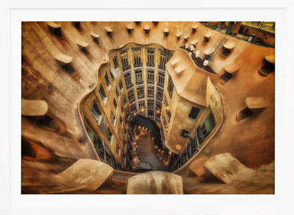 A high-angle, downward view of the inner courtyard of an architecturally unique building, likely Casa Milà by Gaudí. The warm, sandy-colored walls curve and undulate organically, surrounding the multi-story structure with its many windows. The courtyard below is lit, casting a warm glow. Decor