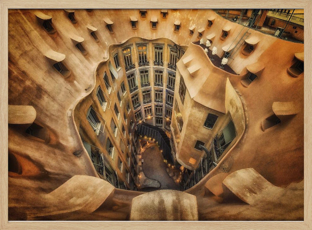 A high-angle, downward view of the inner courtyard of an architecturally unique building, likely Casa Milà by Gaudí. The warm, sandy-colored walls curve and undulate organically, surrounding the multi-story structure with its many windows. The courtyard below is lit, casting a warm glow. Decor