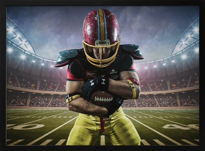 A framed poster of a determined football player in a red and yellow uniform standing on a field in a packed stadium. He is holding a football close to his chest with his head bowed, under bright stadium lights. Decor