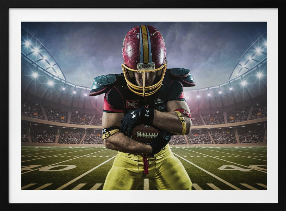 A framed poster of a determined football player in a red and yellow uniform standing on a field in a packed stadium. He is holding a football close to his chest with his head bowed, under bright stadium lights. Decor