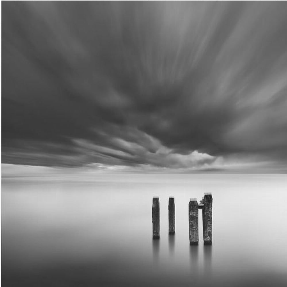 A framed, square, black and white long-exposure photograph of a minimalist seascape. Four dark wooden pilings emerge from still, misty water under a dramatic, streaked sky of dark clouds. Wall Art