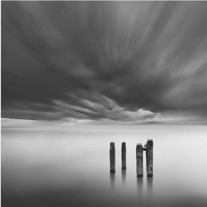 A framed, square, black and white long-exposure photograph of a minimalist seascape. Four dark wooden pilings emerge from still, misty water under a dramatic, streaked sky of dark clouds. Wall Art