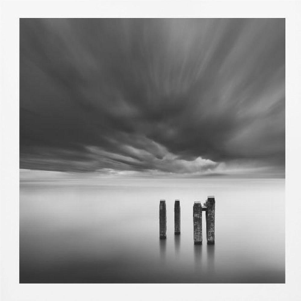 A framed, square, black and white long-exposure photograph of a minimalist seascape. Four dark wooden pilings emerge from still, misty water under a dramatic, streaked sky of dark clouds. Wall Art