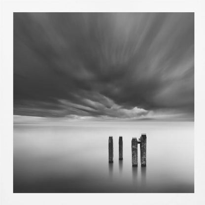 A framed, square, black and white long-exposure photograph of a minimalist seascape. Four dark wooden pilings emerge from still, misty water under a dramatic, streaked sky of dark clouds. Wall Art