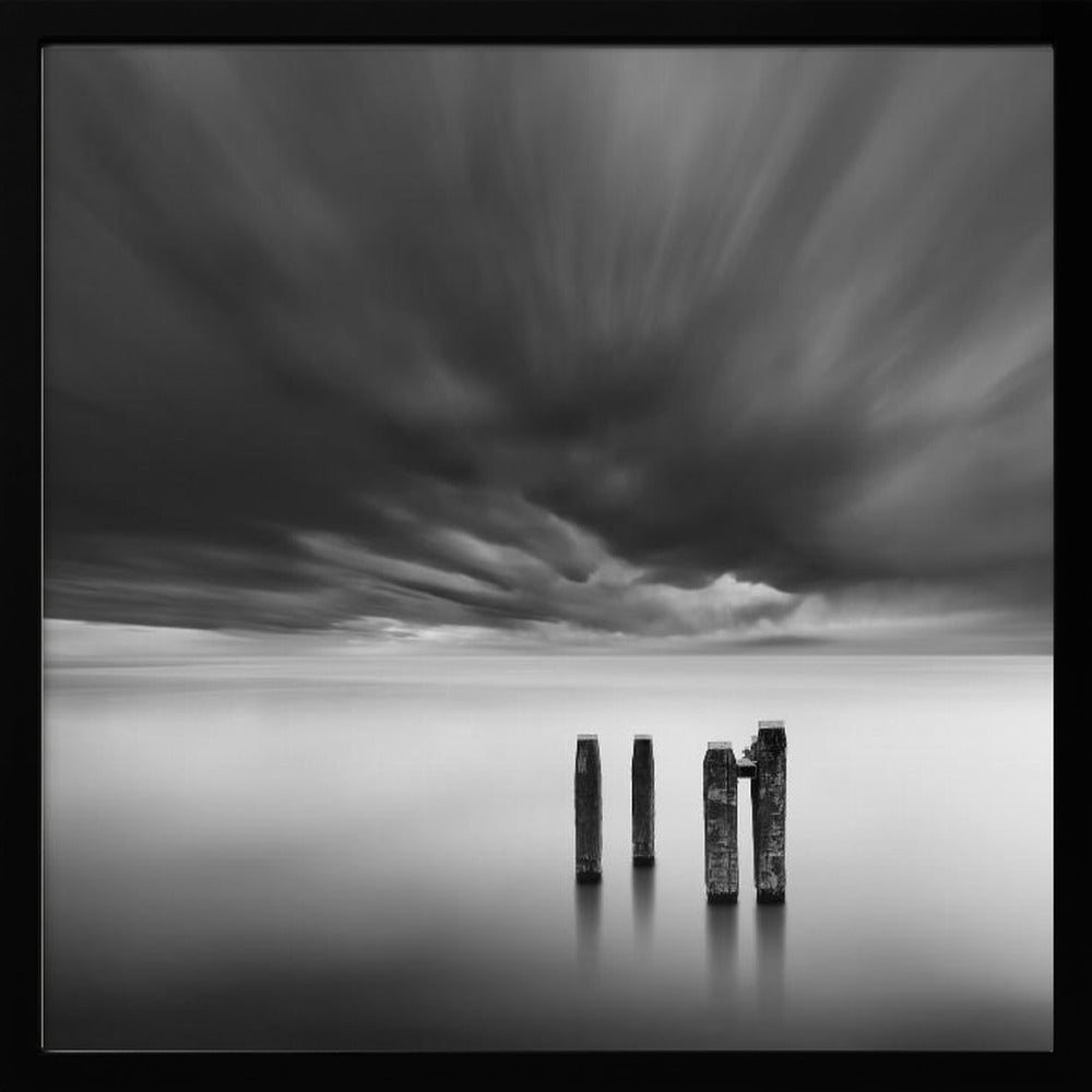 A framed, square, black and white long-exposure photograph of a minimalist seascape. Four dark wooden pilings emerge from still, misty water under a dramatic, streaked sky of dark clouds. Wall Art