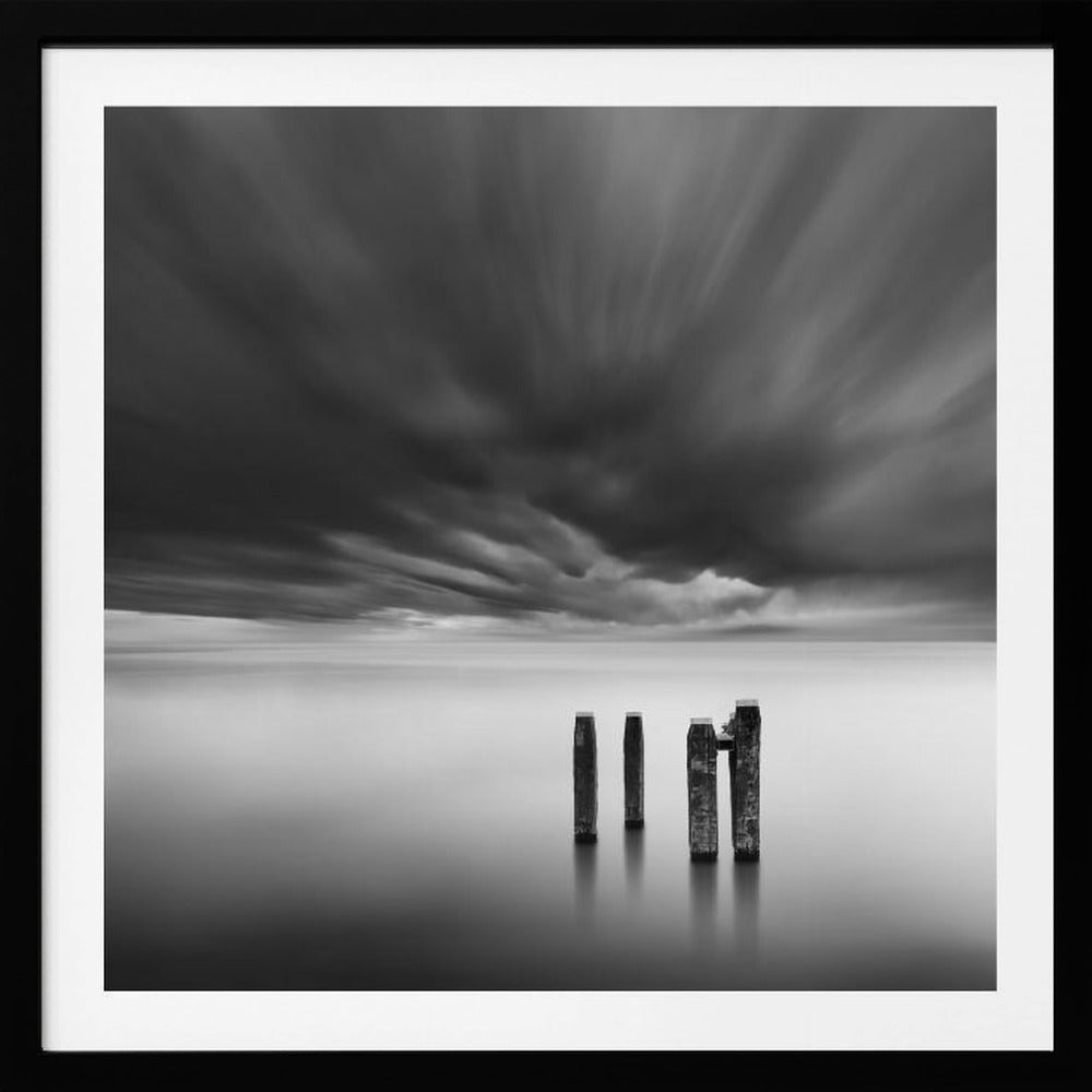 A framed, square, black and white long-exposure photograph of a minimalist seascape. Four dark wooden pilings emerge from still, misty water under a dramatic, streaked sky of dark clouds. Wall Art