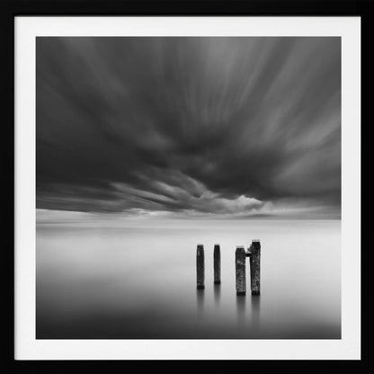 A framed, square, black and white long-exposure photograph of a minimalist seascape. Four dark wooden pilings emerge from still, misty water under a dramatic, streaked sky of dark clouds. Wall Art