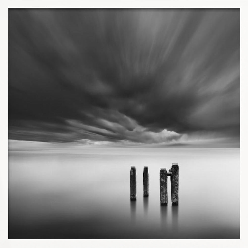 A framed, square, black and white long-exposure photograph of a minimalist seascape. Four dark wooden pilings emerge from still, misty water under a dramatic, streaked sky of dark clouds. Wall Art
