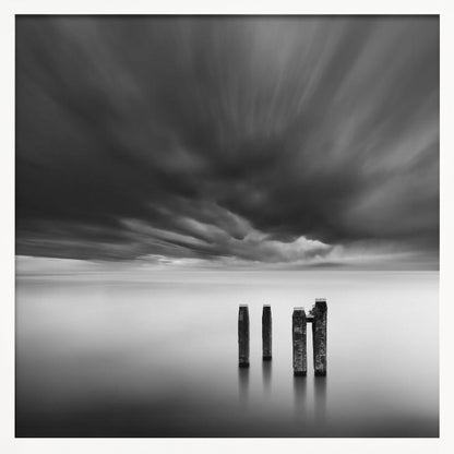 A framed, square, black and white long-exposure photograph of a minimalist seascape. Four dark wooden pilings emerge from still, misty water under a dramatic, streaked sky of dark clouds. Wall Art