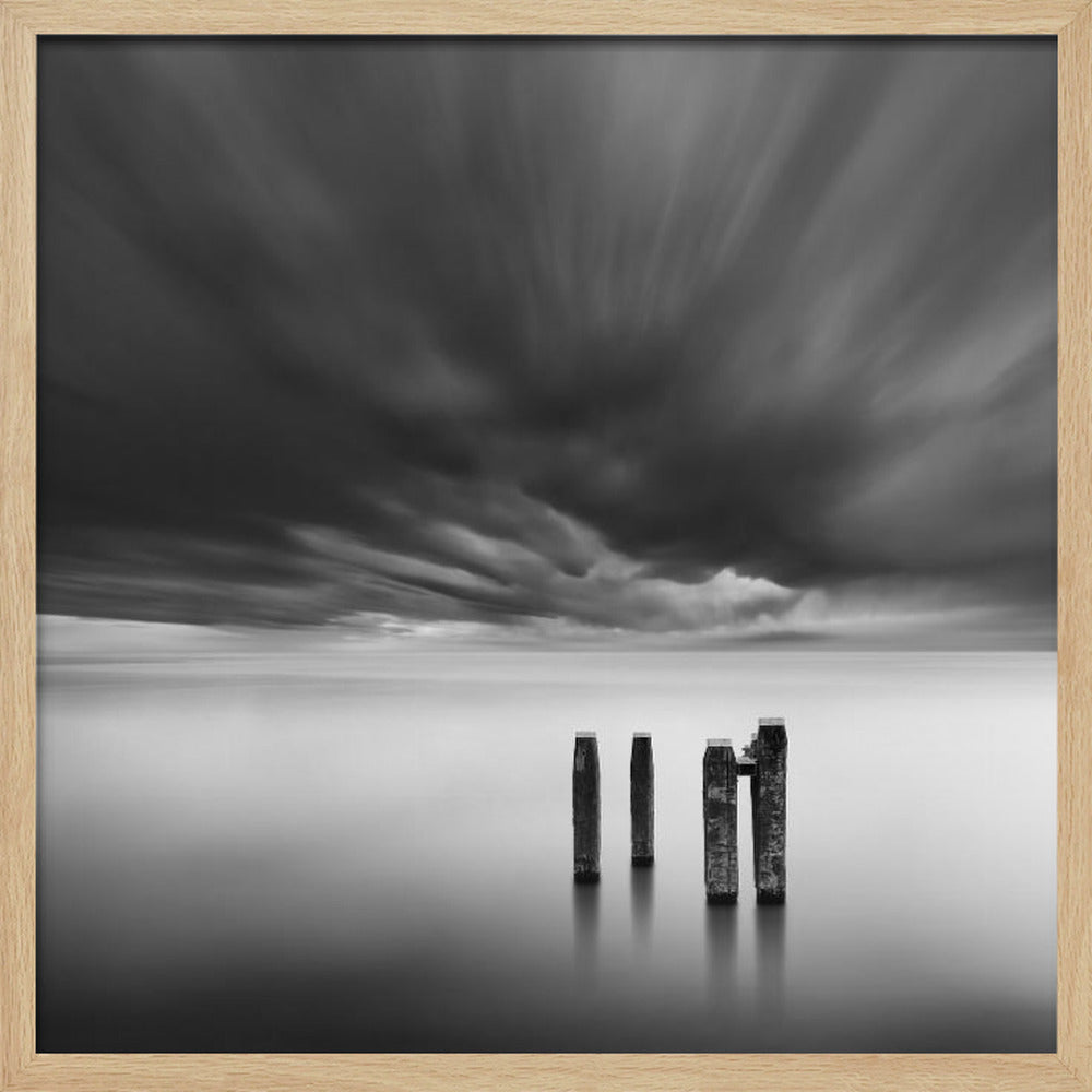 A framed, square, black and white long-exposure photograph of a minimalist seascape. Four dark wooden pilings emerge from still, misty water under a dramatic, streaked sky of dark clouds. Wall Art