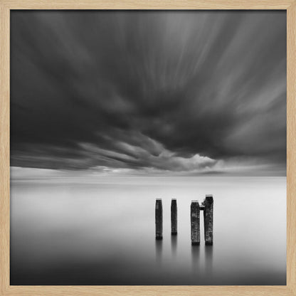 A framed, square, black and white long-exposure photograph of a minimalist seascape. Four dark wooden pilings emerge from still, misty water under a dramatic, streaked sky of dark clouds. Wall Art