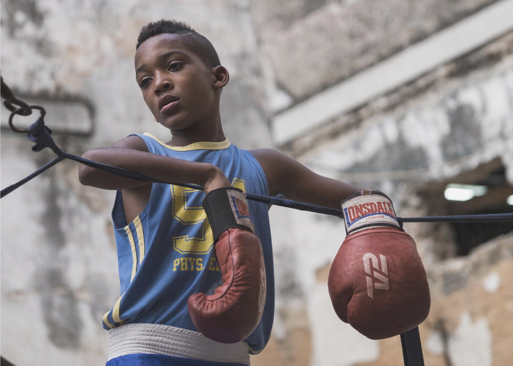 A young Black boy in a blue jersey and red boxing gloves leans pensively against the ropes of a boxing ring, looking away with a thoughtful expression against a blurred, weathered background. Wall Art
