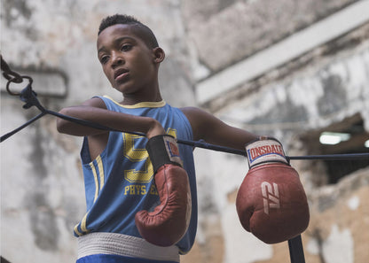 A young Black boy in a blue jersey and red boxing gloves leans pensively against the ropes of a boxing ring, looking away with a thoughtful expression against a blurred, weathered background. Wall Art