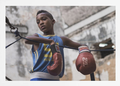 A young Black boy in a blue jersey and red boxing gloves leans pensively against the ropes of a boxing ring, looking away with a thoughtful expression against a blurred, weathered background. Wall Art