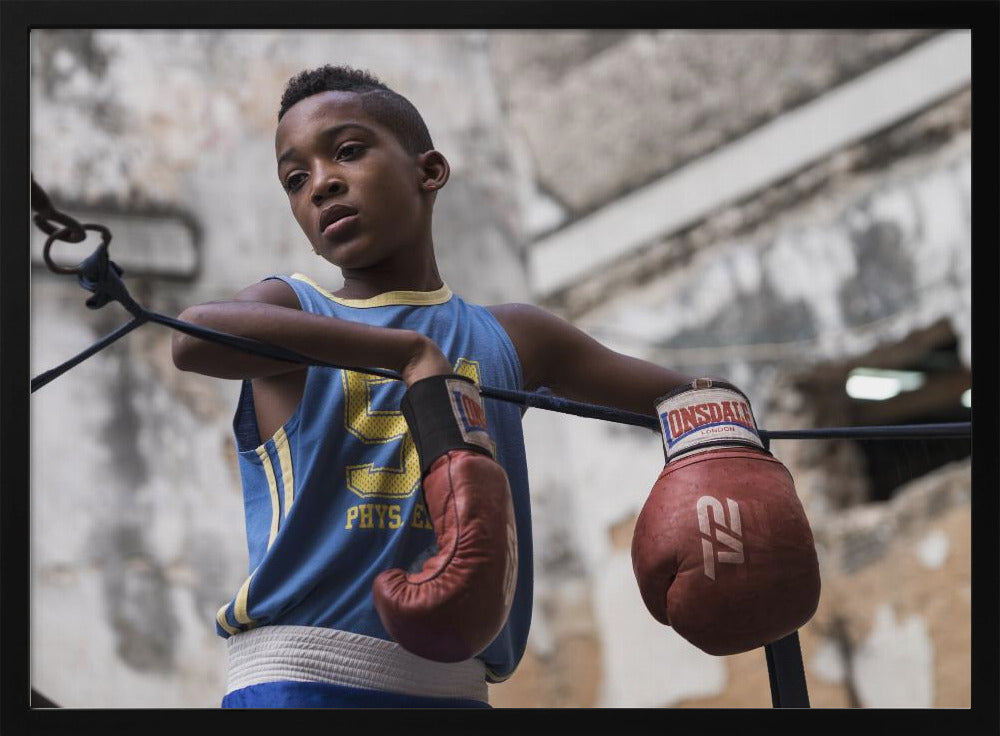 A young Black boy in a blue jersey and red boxing gloves leans pensively against the ropes of a boxing ring, looking away with a thoughtful expression against a blurred, weathered background. Wall Art