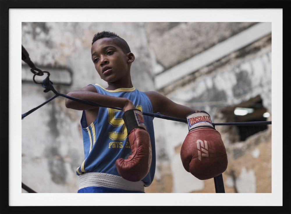 A young Black boy in a blue jersey and red boxing gloves leans pensively against the ropes of a boxing ring, looking away with a thoughtful expression against a blurred, weathered background. Wall Art