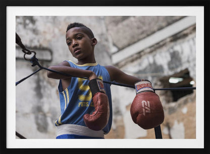 A young Black boy in a blue jersey and red boxing gloves leans pensively against the ropes of a boxing ring, looking away with a thoughtful expression against a blurred, weathered background. Wall Art