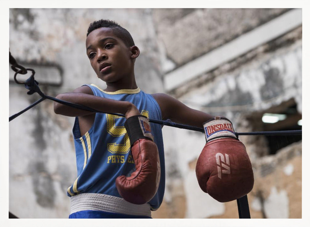 A young Black boy in a blue jersey and red boxing gloves leans pensively against the ropes of a boxing ring, looking away with a thoughtful expression against a blurred, weathered background. Wall Art